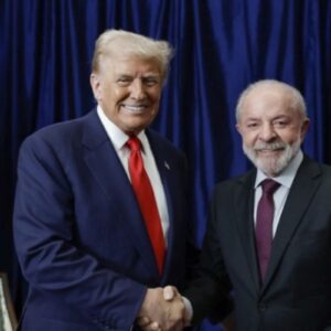 Reunião entre Donald Trump e Luiz Inácio Lula da Silva em evento político, com ambos sorrindo e apertando as mãos, ao fundo bandeira dos Estados Unidos e do Brasil.