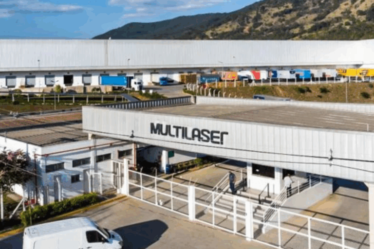 Imagem do centro de distribuição Multlaser com fila de caminhões carregados na frente, localizado em uma região com relevo montanhoso ao fundo, sob céu claro.