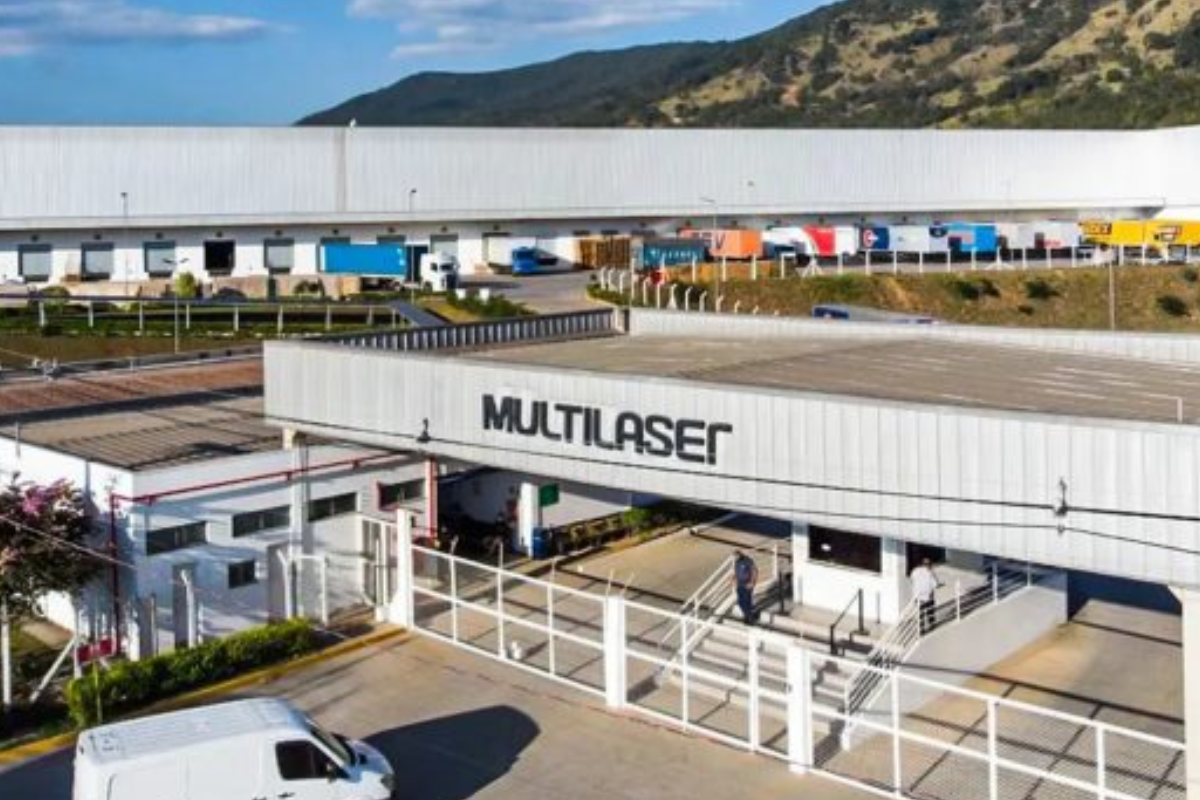 Imagem do centro de distribuição Multlaser com fila de caminhões carregados na frente, localizado em uma região com relevo montanhoso ao fundo, sob céu claro.