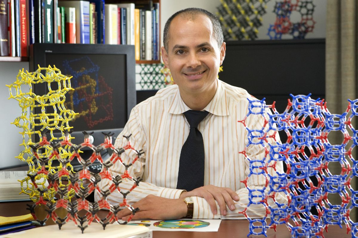 Omar Yaghi com estrutura molecular, cercado por modelos de estruturas químicas coloridas, em um laboratório de pesquisa.