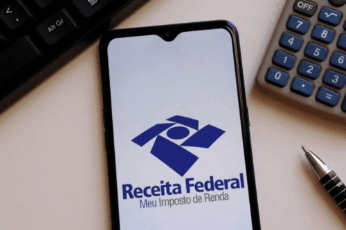 Imagem de um celular exibindo o logo da Receita Federal, símbolo de tributação e impostos, cercado por uma calculadora, caneta e teclado de computador, representando assuntos relacionados à declaração de Imposto de Renda no Brasil.