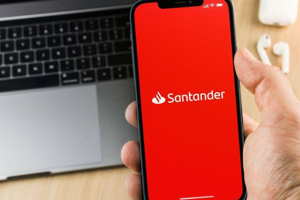 Pessoa segurando um smartphone exibindo o aplicativo do banco Santander, com um teclado de laptop ao fundo.