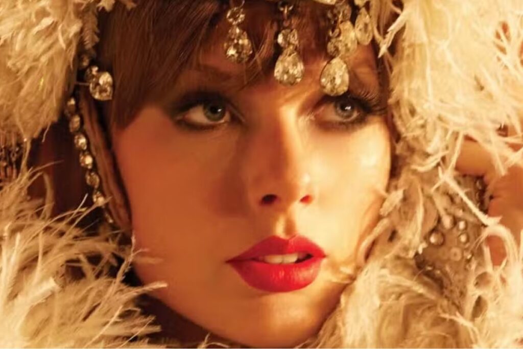 Close-up da Taylor Swift com maquiagem marcante e olhos azuis, usando acessórios de joia brilhantes e roupas elegantes, rodeada de penas.