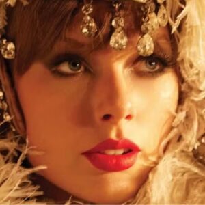 Close-up da Taylor Swift com maquiagem marcante e olhos azuis, usando acessórios de joia brilhantes e roupas elegantes, rodeada de penas.