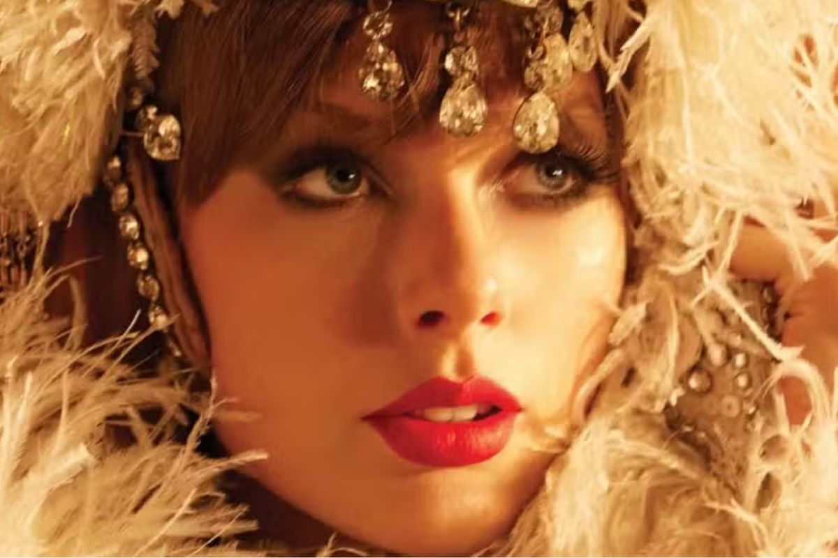 Close-up da Taylor Swift com maquiagem marcante e olhos azuis, usando acessórios de joia brilhantes e roupas elegantes, rodeada de penas.