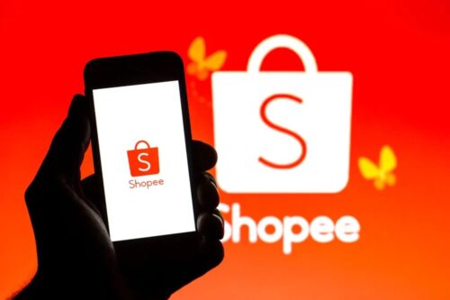 Silhueta de uma mão segurando um smartphone com o aplicativo da Shopee aberto. O logotipo laranja da Shopee (uma sacola de compras com um 'S') é visível na tela e também no fundo em um fundo vermelho brilhante com borboletas laranjas/amarelas.