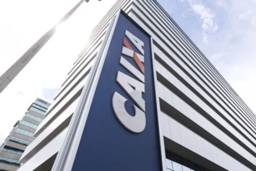 Vista em ângulo de um prédio de escritórios alto e moderno com o logotipo 'CAIXA' em letras grandes e brancas sobre um painel azul marinho.