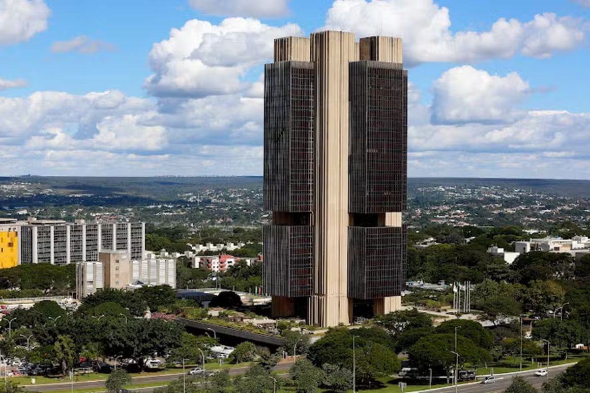 Vista externa do Edifício-Sede do Banco Central do Brasil (BCB) em Brasília, com sua arquitetura brutalista característica, torres escuras verticais e detalhes horizontais claros, sob um céu azul com nuvens brancas, rodeado por uma vasta área verde e a paisagem urbana da capital federal ao fundo.