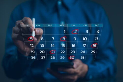 Uma pessoa, em um ambiente de luz azul, está marcando a data '5' (domingo) em um calendário digital flutuante com uma caneta stylus. As datas '2', '5', '8', '18', '21' e '30' estão circuladas, sugerindo o agendamento de eventos ou compromissos importantes ao longo do mês.