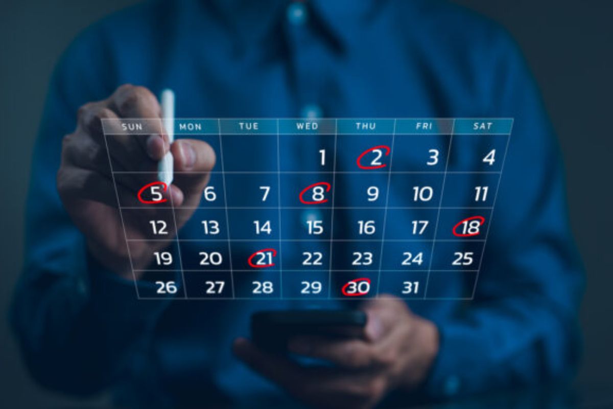 Uma pessoa, em um ambiente de luz azul, está marcando a data '5' (domingo) em um calendário digital flutuante com uma caneta stylus. As datas '2', '5', '8', '18', '21' e '30' estão circuladas, sugerindo o agendamento de eventos ou compromissos importantes ao longo do mês.