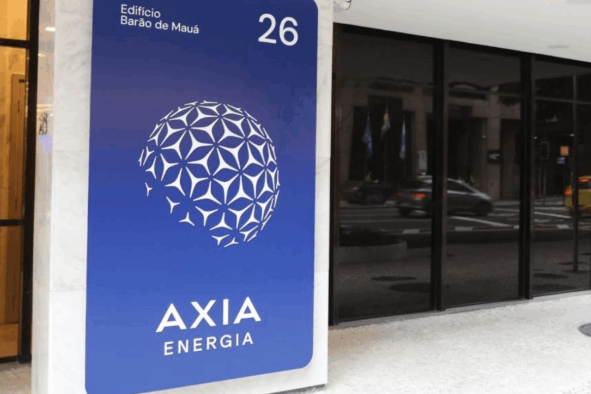 Placa de identificação da empresa Axia Energia fixada em uma coluna de mármore na entrada de um edifício. A placa é azul escura e exibe o logo da empresa (uma esfera geométrica branca), o nome "AXIA ENERGIA" e o endereço "Edifício Barão de Mauá 26" em letras brancas. Ao fundo, vê-se uma fachada de vidro escuro.