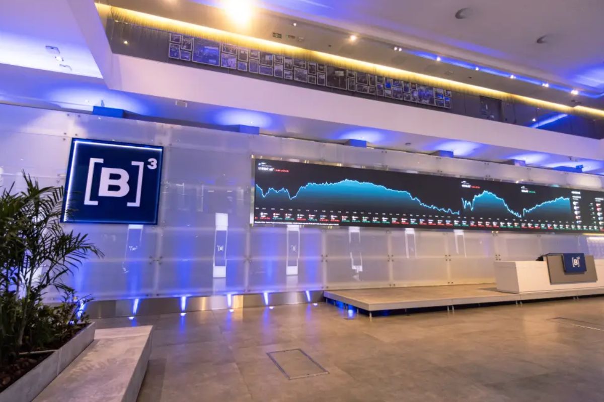 Vista interna moderna do prédio da B3, a bolsa de valores brasileira em São Paulo. O ambiente é iluminado em tons de azul e roxo. No centro, uma tela de LED horizontal e muito longa exibe um gráfico de linha azul mostrando as flutuações do mercado, acompanhado de dados e números. À esquerda da tela, um grande painel luminoso exibe o logotipo da B3: a letra 'B' dentro de um colchete, seguido pelo número '3'.