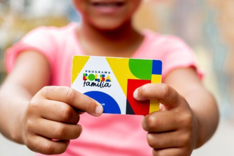 Uma criança, vestindo uma camisa rosa, segura em primeiro plano o cartão de benefícios do Programa Bolsa Família, que apresenta um design colorido e moderno.