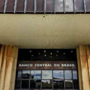 Fachada da entrada principal do Banco Central do Brasil. O nome "BANCO CENTRAL DO BRASIL" está em destaque acima das portas de vidro, sob um teto saliente e cercado por painéis de concreto com textura vertical em tons terrosos.