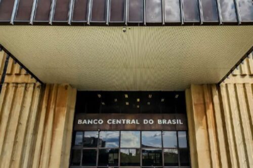 Fachada da entrada principal do Banco Central do Brasil. O nome "BANCO CENTRAL DO BRASIL" está em destaque acima das portas de vidro, sob um teto saliente e cercado por painéis de concreto com textura vertical em tons terrosos.