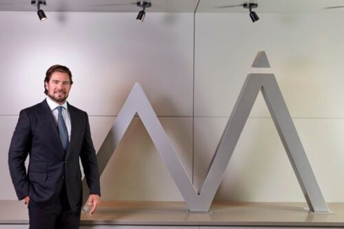 Homem branco, de barba, vestindo um terno escuro com gravata, em pé ao lado de um grande logotipo escultural cinza que se assemelha a um pico de montanha com um triângulo no topo.