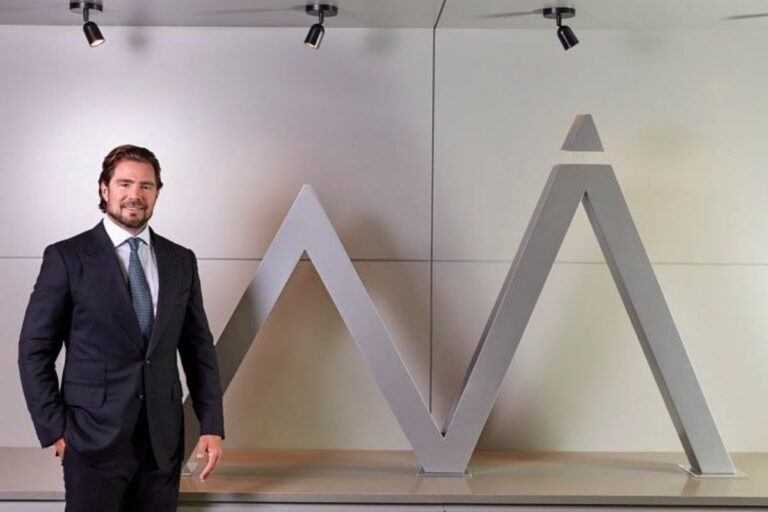 Homem branco, de barba, vestindo um terno escuro com gravata, em pé ao lado de um grande logotipo escultural cinza que se assemelha a um pico de montanha com um triângulo no topo.