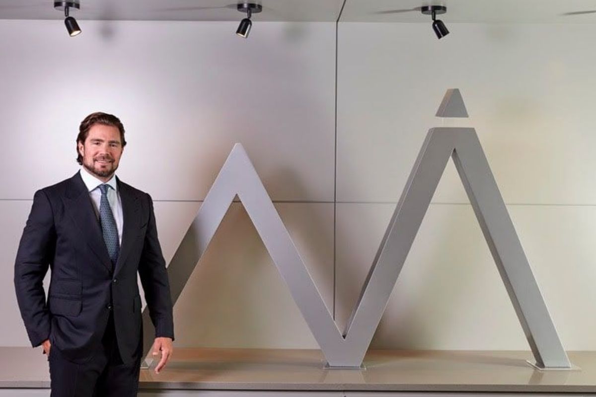 Homem branco, de barba, vestindo um terno escuro com gravata, em pé ao lado de um grande logotipo escultural cinza que se assemelha a um pico de montanha com um triângulo no topo.