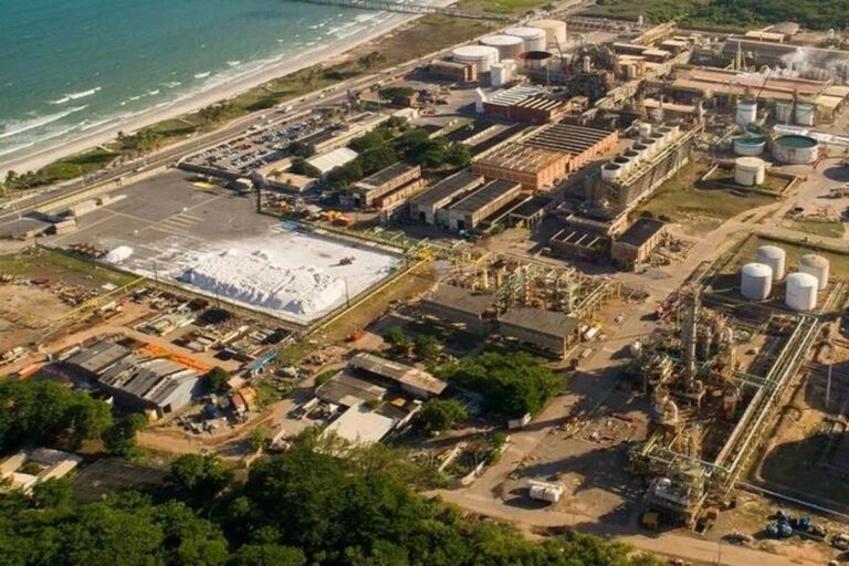 Vista aérea de uma grande planta petroquímica ou industrial (Braskem) localizada à beira-mar. A imagem mostra diversos edifícios industriais, tanques de armazenamento cilíndricos, tubulações e uma grande pilha de material branco (possivelmente sal ou produto químico) em uma área de pátio. No canto superior esquerdo, o oceano e a praia são visíveis.