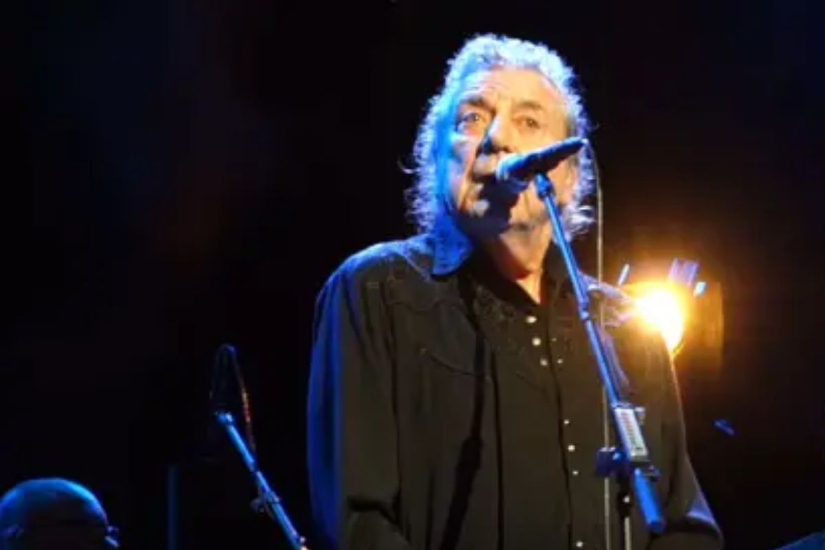 O cantor Robert Plant, do Led Zeppelin, em close-up no palco, cantando em um microfone. Ele tem cabelos grisalhos e longos, veste uma camisa preta e está sob luzes de show, com uma forte luz amarela iluminando a cena por trás. A imagem é de uma apresentação histórica do clássico 'Kashmir' em Londres.