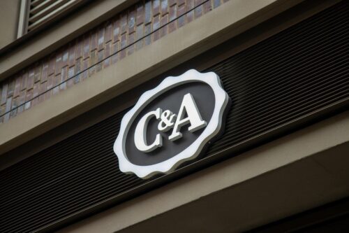 O logo da C&A, uma varejista de moda internacional, exibido em um letreiro circular e ondulado, com as letras 'C&A' em branco sobre um fundo oval escuro, fixado na fachada de um edifício.