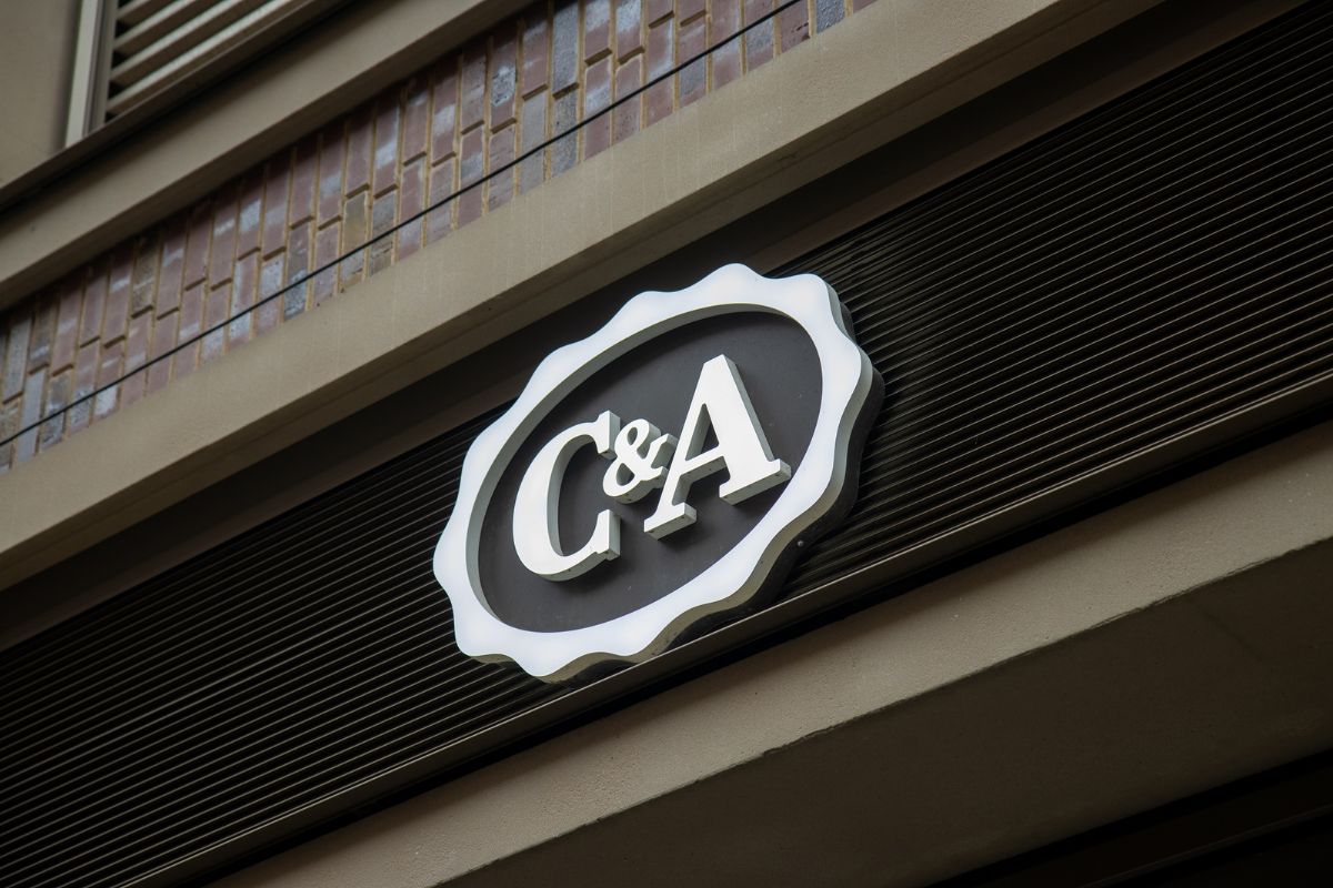 O logo da C&A, uma varejista de moda internacional, exibido em um letreiro circular e ondulado, com as letras 'C&A' em branco sobre um fundo oval escuro, fixado na fachada de um edifício.