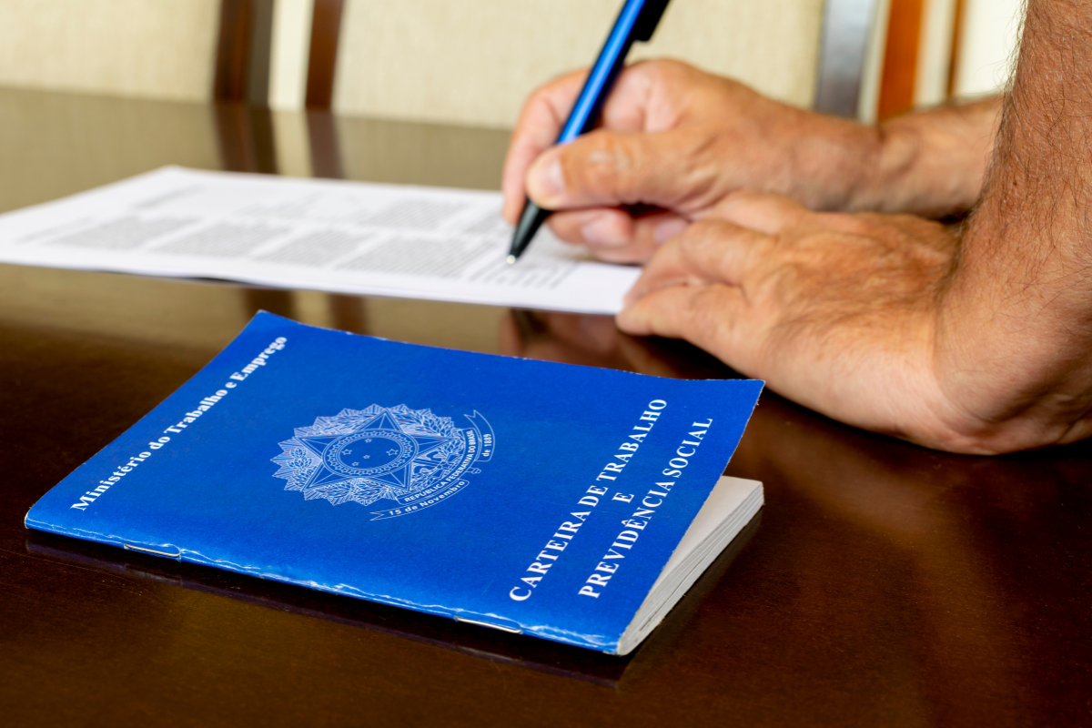 Pessoa assinando a Carteira de Trabalho e Previdência Social ao lado de um documento, possivelmente um formulário ou papel de registros.