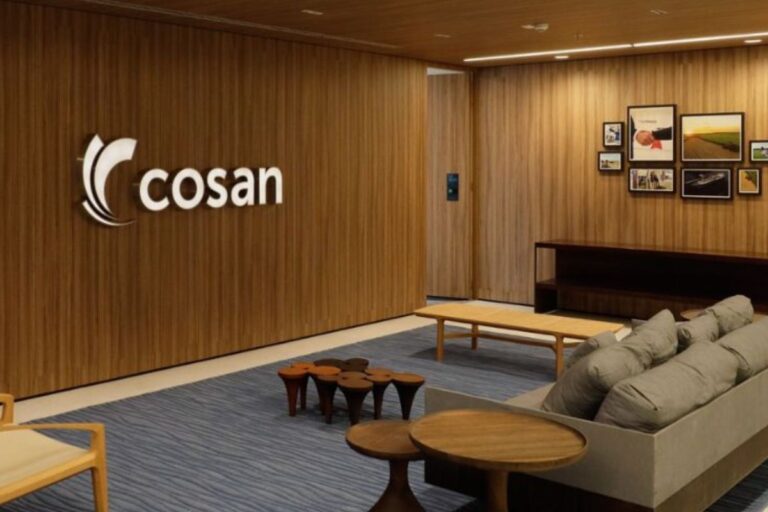 Interior moderno e elegante de uma recepção ou sala de espera da Cosan, com o logotipo da empresa em relevo na parede de madeira, um tapete azul e sofás e mesas de centro em madeira e tons neutros.