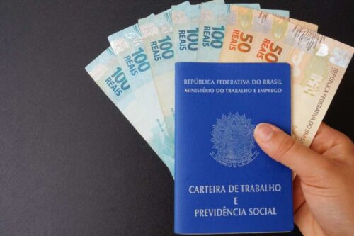 Uma mão segura a capa azul da Carteira de Trabalho e Previdência Social (CTPS) brasileira, colocada sobre uma pilha de notas de Real (R$ 50 e R$ 100), simbolizando trabalho, direitos e pagamento.