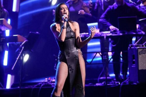 Uma foto de Dua Lipa se apresentando em um palco, cantando em um microfone. Ela está vestindo um longo vestido preto de couro, com cortes laterais de malha arrastão e detalhes de cruzes, e botas pretas de cano alto.