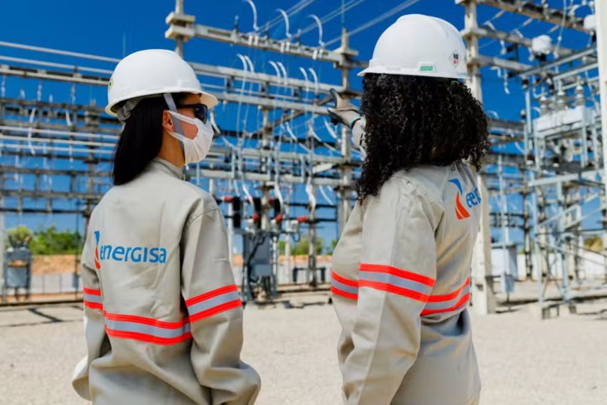 Duas engenheiras ou técnicas, vestindo uniformes da Energisa com faixas reflexivas e capacetes de segurança brancos, estão em uma subestação de energia elétrica.