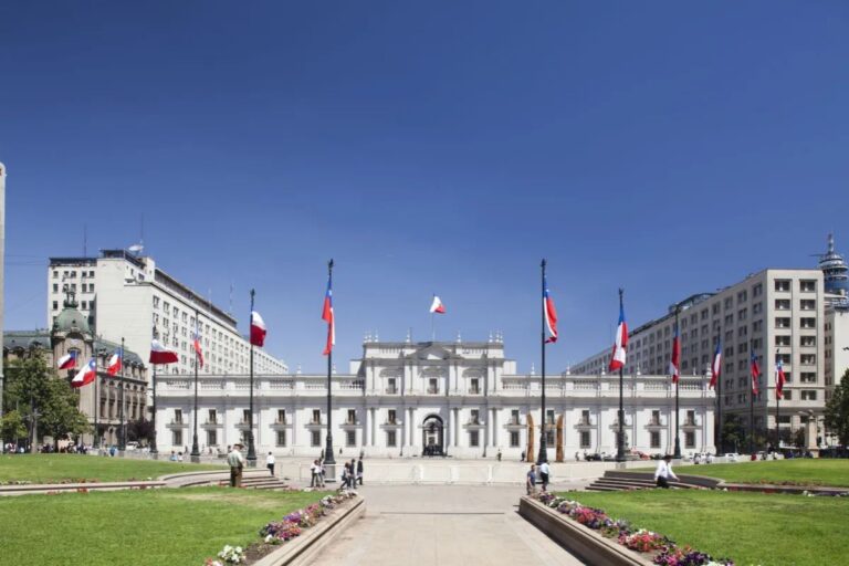 Vista frontal externa do Palácio de La Moneda, a sede do governo chileno, em Santiago. O prédio neoclássico branco está sob um céu azul claro, cercado por uma praça gramada e caminhos pavimentados. Diversas bandeiras do Chile estão hasteadas em postes ao redor da praça e em frente ao palácio.