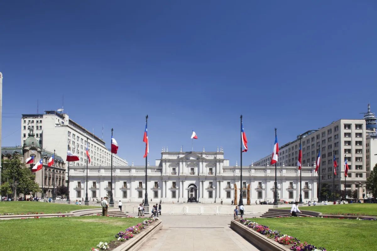 Vista frontal externa do Palácio de La Moneda, a sede do governo chileno, em Santiago. O prédio neoclássico branco está sob um céu azul claro, cercado por uma praça gramada e caminhos pavimentados. Diversas bandeiras do Chile estão hasteadas em postes ao redor da praça e em frente ao palácio.