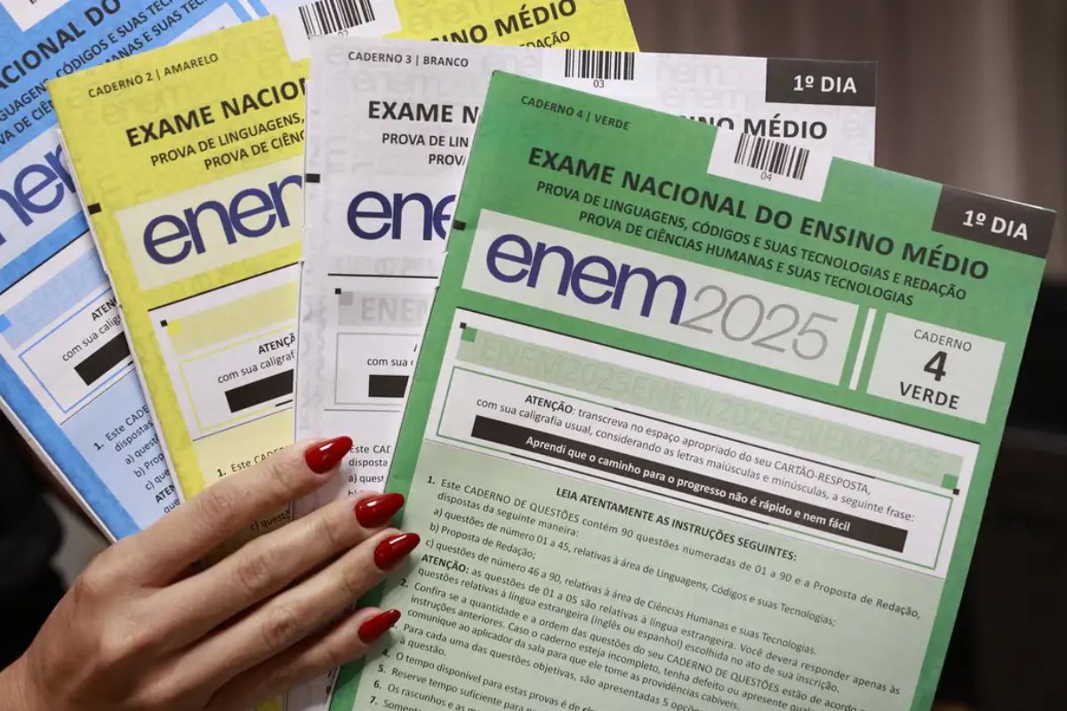 Quatro cadernos de prova do Exame Nacional do Ensino Médio (ENEM) de 2025 estão empilhados e parcialmente visíveis, sendo segurados por uma mão com unhas pintadas de vermelho.