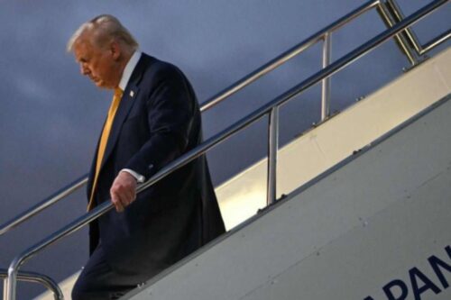 O presidente dos Estados Unidos, Donald Trump, usando um terno escuro e uma gravata amarela, desce a escada de um avião. Ele está de perfil, olhando para baixo, segurando o corrimão prateado da escada com a mão direita.