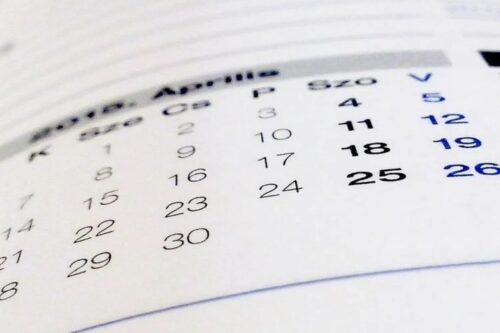 Uma visão em close-up e desfocada de um calendário em papel, mostrando os dias e números de um mês, provavelmente 'Abril' ou 'Aprilie', com os números 18, 19 e 25 destacados em negrito.