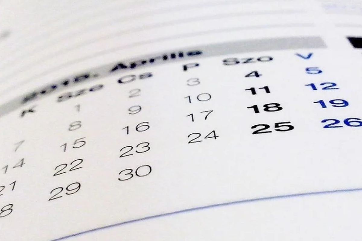 Uma visão em close-up e desfocada de um calendário em papel, mostrando os dias e números de um mês, provavelmente 'Abril' ou 'Aprilie', com os números 18, 19 e 25 destacados em negrito.