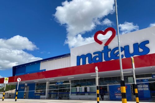 Fachada externa e de baixo ângulo de uma nova loja do Grupo Mateus, com a logomarca azul e um coração vermelho no topo, sob um céu azul com nuvens brancas.