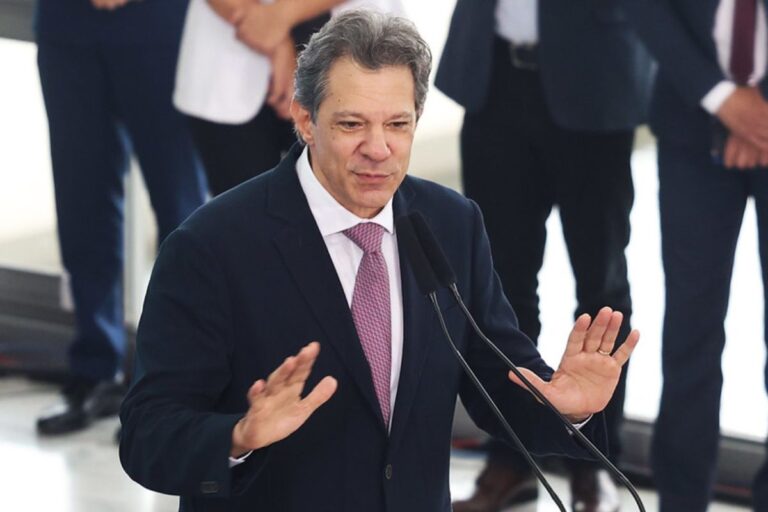 O Ministro da Fazenda, Fernando Haddad, de terno azul-marinho e gravata rosa com padrão, fala em um púlpito com microfones duplos, gesticulando com as mãos abertas.