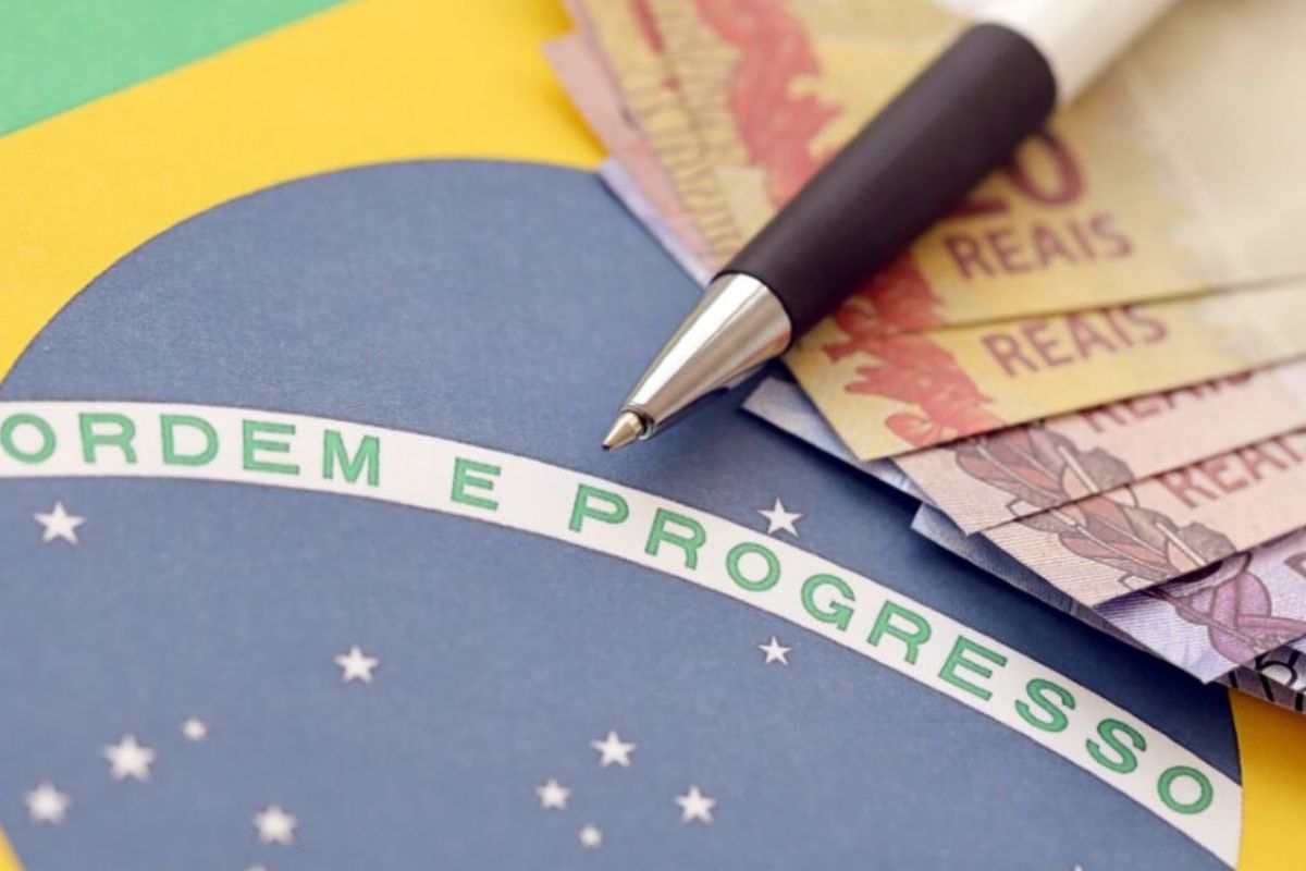 Uma imagem em close-up mostrando a faixa 'ORDEM E PROGRESSO' da bandeira do Brasil. Sobre a bandeira, estão algumas notas de Real brasileiro e uma caneta preta e prateada, com a ponta apontando para a faixa.