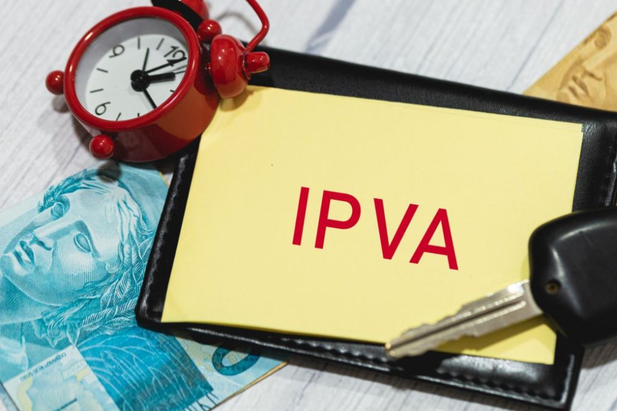 Composição de uma carteira ou porta-documentos preta, uma nota de Real, uma chave de carro, um pequeno despertador vermelho (simbolizando prazo) e um papel amarelo com a inscrição "IPVA" em letras vermelhas.