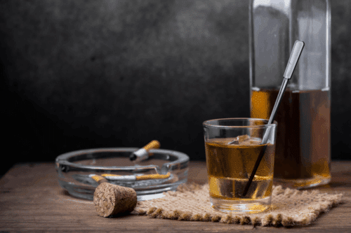 Cigarros, cinzeiro com pontas apagadas, bebida alcoólica em copo com canudo e garrafa de aguardente na mesa, ambiente com fundo escuro