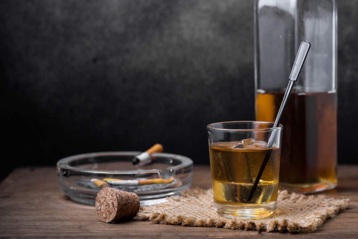 Cigarros, cinzeiro com pontas apagadas, bebida alcoólica em copo com canudo e garrafa de aguardente na mesa, ambiente com fundo escuro