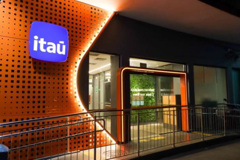 Fachada noturna de uma agência bancária do Itaú com iluminação laranja vibrante. O logotipo azul-claro do Itaú está fixado em uma parede laranja texturizada com pequenos quadrados.