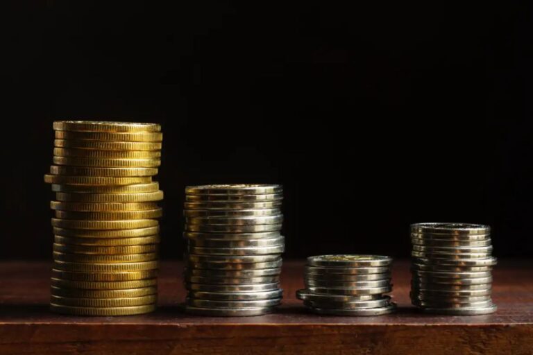 Quatro pilhas de moedas, variando em altura e cor (algumas douradas e outras prateadas), dispostas em uma superfície de madeira escura contra um fundo preto.