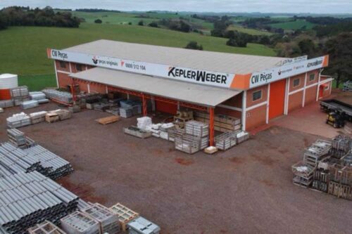 Vista aérea ou elevada do armazém de peças e depósito da Kepler Weber. O prédio de tijolos vermelhos e teto cinza exibe o logotipo "KEPLER WEBER" em destaque. O pátio em frente está repleto de paletes, caixas e tubos de PVC estocados, com um caminhão visível à direita. Ao fundo, uma paisagem rural com colinas verdes.