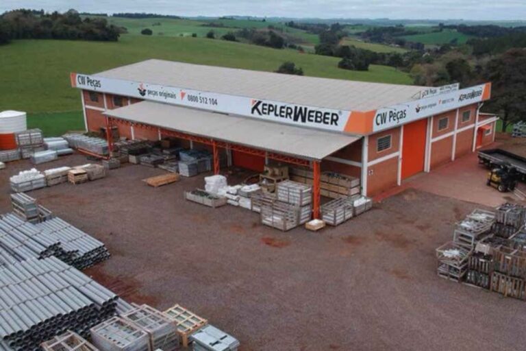 Vista aérea ou elevada do armazém de peças e depósito da Kepler Weber. O prédio de tijolos vermelhos e teto cinza exibe o logotipo "KEPLER WEBER" em destaque. O pátio em frente está repleto de paletes, caixas e tubos de PVC estocados, com um caminhão visível à direita. Ao fundo, uma paisagem rural com colinas verdes.