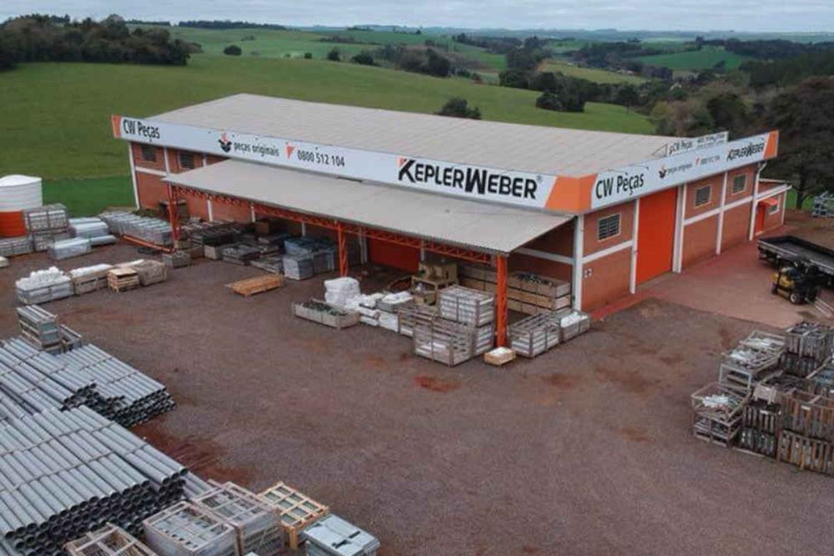 Vista aérea ou elevada do armazém de peças e depósito da Kepler Weber. O prédio de tijolos vermelhos e teto cinza exibe o logotipo "KEPLER WEBER" em destaque. O pátio em frente está repleto de paletes, caixas e tubos de PVC estocados, com um caminhão visível à direita. Ao fundo, uma paisagem rural com colinas verdes.