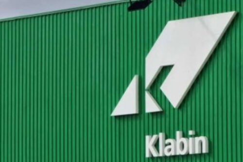 O logotipo e o nome 'Klabin' em branco, em relevo, fixados em uma parede externa verde e canelada de um prédio industrial ou fábrica.