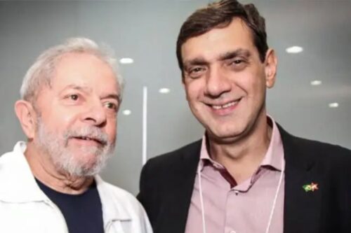 O presidente Luiz Inácio Lula da Silva, de barba branca e camisa polo branca, posa ao lado de seu filho Fábio Luís Lula da Silva ('Lulinha'), que veste um blazer preto sobre uma camisa rosa.